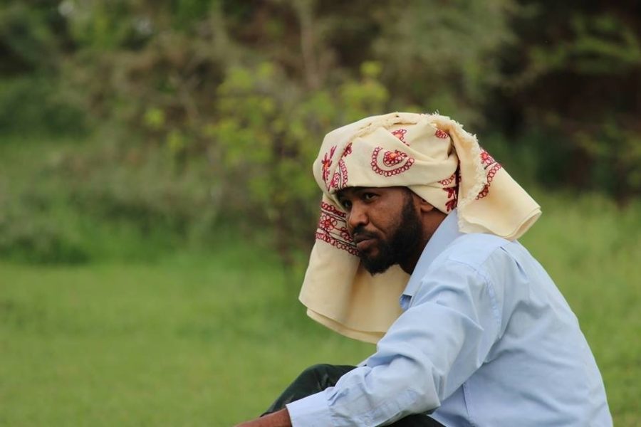 Weedhsame, a male Somali poet, sits wearing a blue shirt in nature, wearing a headscarf